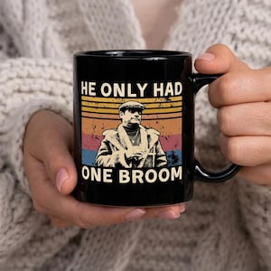 May include: Black ceramic mug featuring a retro design with the text "HE ONLY HAD ONE BROOM" and an image of a man in a coat and cap. The design has a vintage color scheme of orange, yellow, red, and blue.