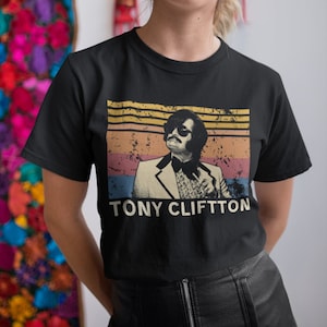Puede incluir: Camiseta negra con una imagen de estilo vintage de Tony Clifton, cantante y comediante, con un fondo de puesta de sol retro. El texto "TONY CLIFTON" está impreso debajo de la imagen.