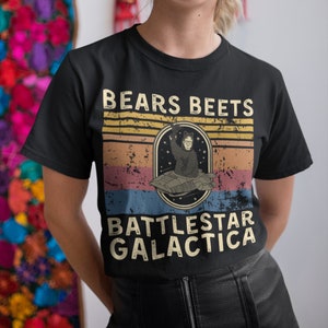 May include: A black t-shirt with a vintage-style graphic featuring a person in a spacesuit with the text "Bears Beets Battlestar Galactica".