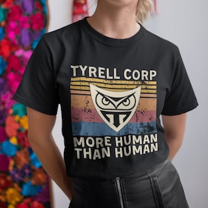 May include: Black t-shirt featuring the text "TYRELL CORP" above a shield-shaped logo with an owl design. Below the logo, the text reads "MORE HUMAN THAN HUMAN". The design has a vintage, distressed look with a retro color scheme.