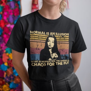 May include: Black t-shirt with a vintage-style graphic featuring a woman with dark hair and a quote that reads "Normal is an illusion. What is normal for the spider is chaos for the fly."