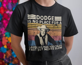 Può includere: Una maglietta nera con una grafica in stile vintage che presenta un uomo con un cappello da cowboy e il testo "Dodge is no place for a gambling man who talks fast but moves slow".