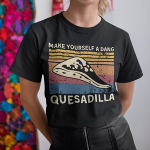 Puede incluir: Camiseta negra con un gráfico de estilo vintage que dice "Hazte una quesadilla". El gráfico presenta una quesadilla con queso derretido y un fondo de puesta de sol de estilo retro.