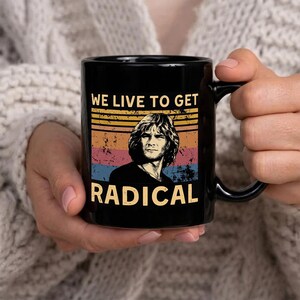 Peut inclure: Mug en céramique noire avec un graphisme de style vintage. Le design comprend un portrait d'un homme aux cheveux longs, sur fond de rayures horizontales jaunes, oranges, roses et bleues. Le texte "WE LIVE TO GET RADICAL" est affiché.