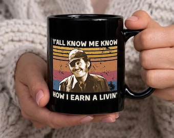 Może przedstawiać: Czarny ceramiczny kubek z retro wzorem. Przedstawia uśmiechniętego mężczyznę w czapce i biały napis "Y'all know me know how I earn a livin'". Tło ma poziome paski w kolorach pomarańczowym, żółtym, różowym i niebieskim.