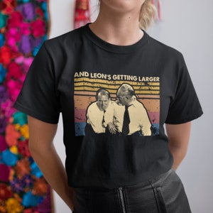 May include: A black t-shirt with a vintage-style graphic of two men. The graphic features a sunset background with the text "AND LEON'S GETTING LARGER".
