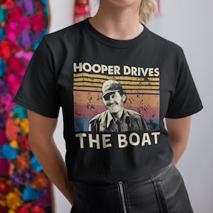 May include: A black t-shirt featuring a vintage-style graphic of a man wearing a hat and the text "HOOPER DRIVES THE BOAT".
