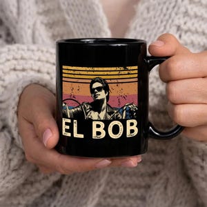 May include: Black ceramic mug featuring a retro design with the text "EL BOB". The design includes a man wearing sunglasses and a leather jacket, set against a backdrop of horizontal stripes in orange, yellow, and pink.