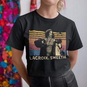 Puede incluir: Una camiseta negra con un gráfico de estilo vintage de una mujer con un atuendo glamuroso, con el texto "LACROIX, SWEETIE" debajo de la imagen.