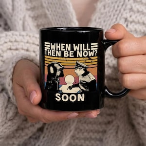 Peut inclure: Mug en céramique noire avec un graphisme rétro. Le texte "WHEN WILL THEN BE NOW?" est au-dessus d'une image de deux personnages et le mot "SOON" en dessous. La tasse a une anse et un intérieur noirs.