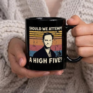 Pode incluir: Caneca de cerâmica preta com o texto "SHOULD WE ATTEMPT A HIGH FIVE?" e um retrato de um homem. O design inclui um bloco de cores retrô laranja, amarelo, azul e vermelho. Um presente divertido.