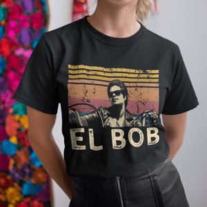 Könnte beinhalten: Schwarzes T-Shirt mit einem Vintage-Grafikdruck eines Mannes, der ein Motorrad fährt. Die Grafik enthält einen Sonnenuntergangs-Hintergrund mit dem Text "EL BOB" in Weiß.