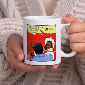 May include: White ceramic mug featuring a comic strip illustration. The comic shows two figures, one in a cowboy hat, and the text bubbles read "WHAT DID THE FIVE FINGERS SAY TO THE FACE?" and "SLAP!"