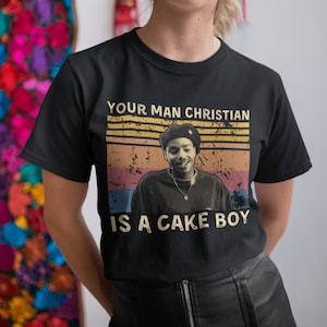 May include: Black t-shirt with a vintage-style graphic of a person wearing a hat and the text "Your Man Christian Is A Cake Boy".