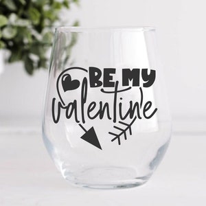 May include: A clear glass wine glass with a black vinyl decal that says "Be My Valentine" with a heart and arrow design.