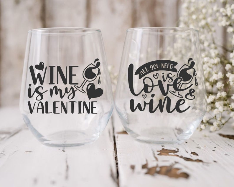 May include: Two clear glass wine glasses with black vinyl lettering. The first glass says "Wine is my Valentine". The second glass says "All you need is love & wine".