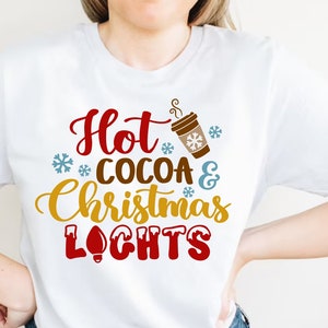 May include: White t-shirt with the text "Hot Cocoa & Christmas Lights" in red, brown, gold, and blue lettering. A stylized image of a cup of hot cocoa with steam coming from it is above the text.