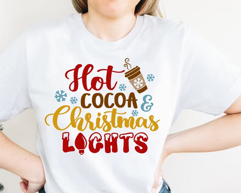 May include: White t-shirt with the text "Hot Cocoa & Christmas Lights" in red, brown, gold, and blue lettering. A stylized image of a cup of hot cocoa with steam coming from it is above the text.