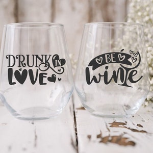 May include: Two clear glass wine glasses with black vinyl decals. The glass on the left says "Drunk in Love" with hearts. The glass on the right says "Be Wine" with hearts.