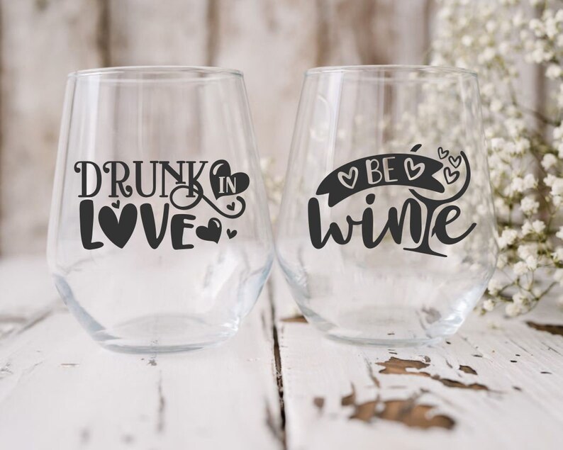 May include: Two clear glass wine glasses with black vinyl decals. The glass on the left says "Drunk in Love" with hearts. The glass on the right says "Be Wine" with hearts.