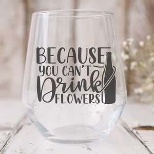 May include: A clear glass wine glass with a black vinyl decal that reads "Because you can't drink flowers". A black silhouette of a wine bottle is next to the text.