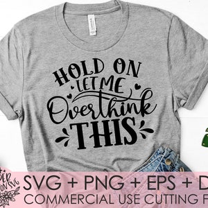 Puede incluir: Una camiseta gris con texto negro que dice "Hold on let me overthink this". El texto est&aacute; en una fuente de letra manuscrita. La camiseta est&aacute; sobre una superficie blanca con un par de pantalones vaqueros azules y una hoja verde.