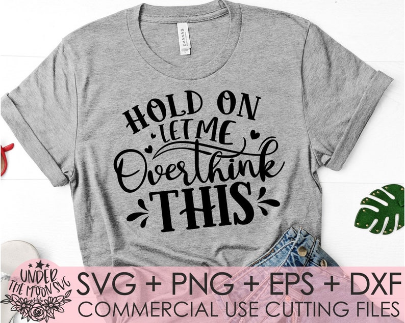 Puede incluir: Una camiseta gris con texto negro que dice "Hold on let me overthink this". El texto est&aacute; en una fuente de letra manuscrita. La camiseta est&aacute; sobre una superficie blanca con un par de pantalones vaqueros azules y una hoja verde.