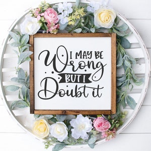 Puede incluir: Un letrero de madera blanco con marco marr&oacute;n, con el texto "I may be wrong but I doubt it" en letras cursivas negras. El letrero est&aacute; rodeado de una corona de flores rosas, blancas y amarillas.