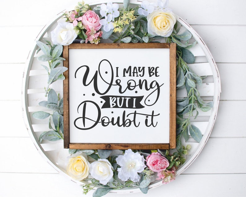 Puede incluir: Un letrero de madera blanco con marco marr&oacute;n, con el texto "I may be wrong but I doubt it" en letras cursivas negras. El letrero est&aacute; rodeado de una corona de flores rosas, blancas y amarillas.