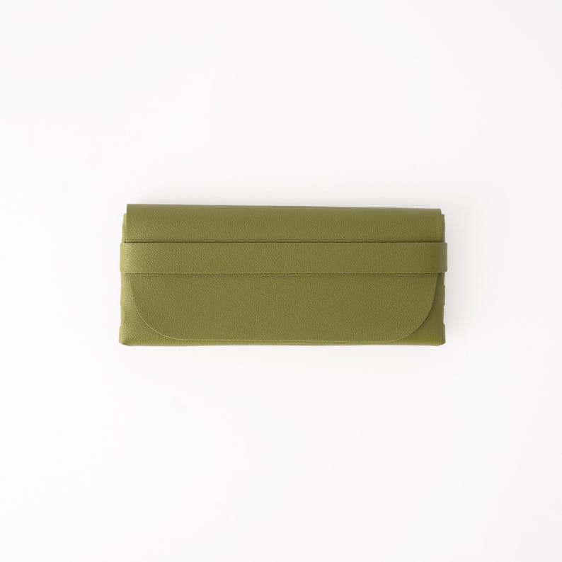 May include: Olive green clutch wallet with a wraparound strap closure. The wallet has a curved edge and a rectangular shape. The material appears to be leather or a leather-like material. The background is white.