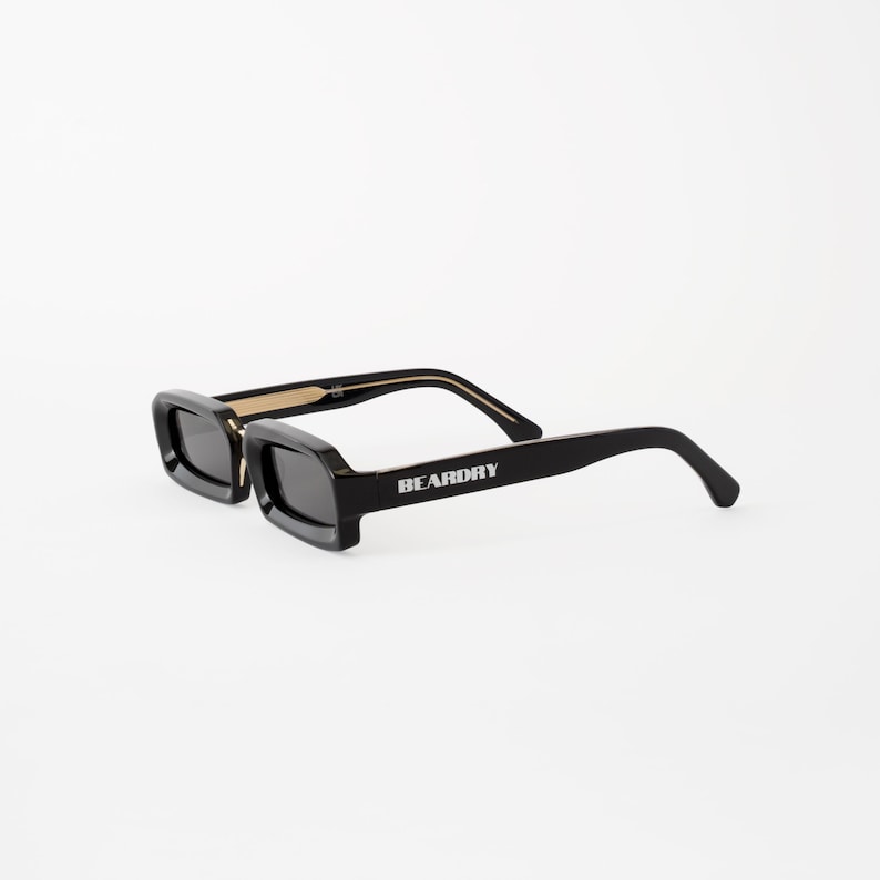 May include: Black rectangular sunglasses with dark lenses. The sunglasses have a thick frame and the text "BEARDRY" is printed on the right arm. The arms are black with gold accents near the hinges. The sunglasses are set against a white background.