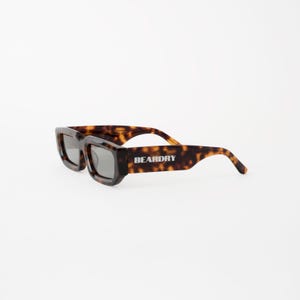 Peut inclure: Lunettes de soleil à motif écaille de tortue avec monture carrée et verres foncés. Le texte "BEARDRY" est imprimé sur la branche droite. Les montures sont marron et noires.