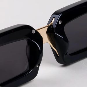 May include: Close-up of black sunglasses with a gold-colored bridge. The sunglasses have a rectangular shape and dark lenses. The frame is thick and glossy, with small silver rivets. The design is modern and sleek.