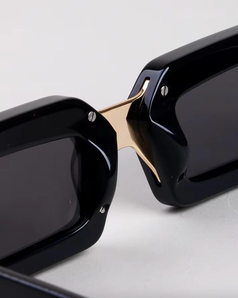 May include: Close-up of black sunglasses with a gold-colored bridge. The sunglasses have a rectangular shape and dark lenses. The frame is thick and glossy, with small silver rivets. The design is modern and sleek.