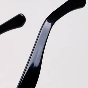 May include: Close-up of the temple arm of black eyeglasses. The arm curves gently and has a glossy finish. The image is set against a plain white background, highlighting the sleek design of the eyewear.