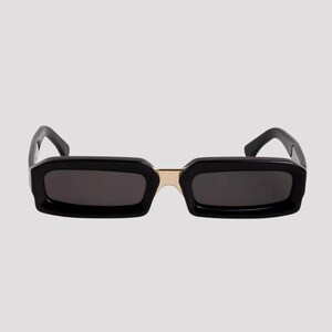 May include: Black rectangular sunglasses with dark lenses and a gold-colored bridge. The frames are thick and angular, giving a bold, modern look. The sunglasses are set against a plain, light background.