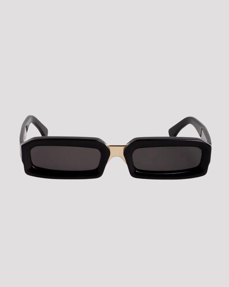 May include: Black rectangular sunglasses with dark lenses and a gold-colored bridge. The frames are thick and angular, giving a bold, modern look. The sunglasses are set against a plain, light background.