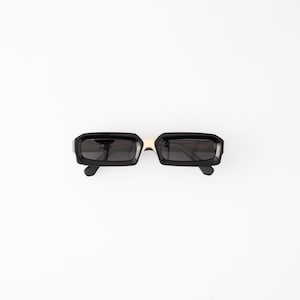 May include: Black rectangular sunglasses with a gold-colored bridge. The sunglasses have a thick, glossy black frame and dark lenses. The sunglasses are set against a white background.