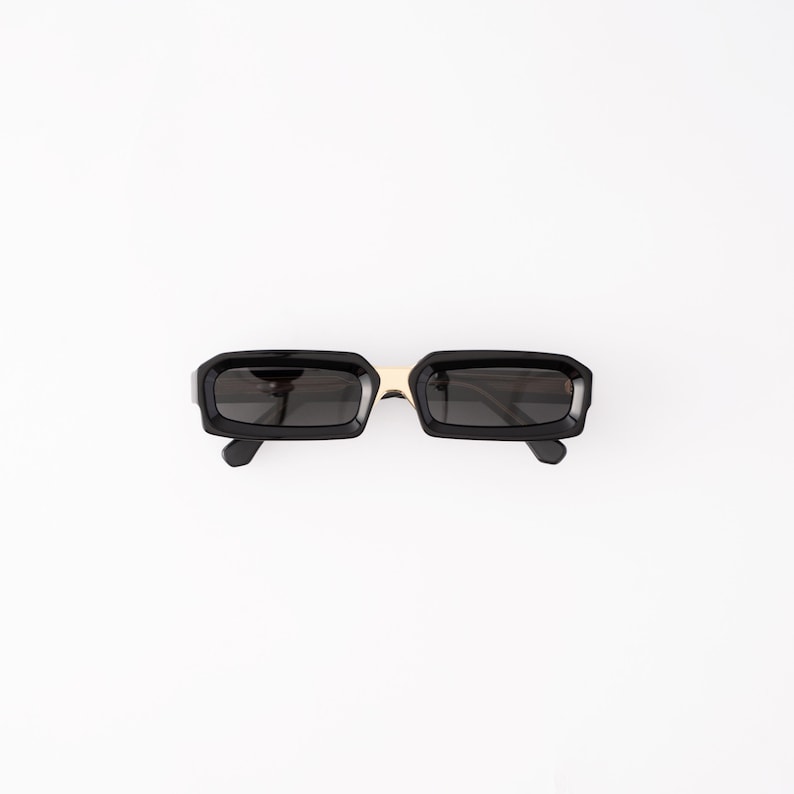 May include: Black rectangular sunglasses with a gold-colored bridge. The sunglasses have a thick, glossy black frame and dark lenses. The sunglasses are set against a white background.
