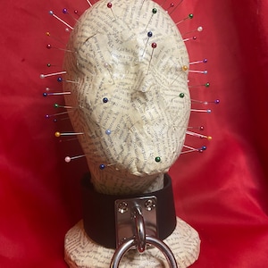 Può includere: Una testa di manichino ricoperta da un collage di pagine di libro con spilli colorati inseriti nella carta. La testa indossa un collare di pelle nera con un anello d'argento.