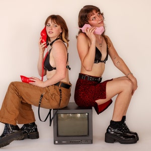 May include: Two people are seated, each holding a vintage telephone. One holds a red phone, the other a pink one. They are wearing black tops and boots. One wears brown pants, the other a red skirt. A vintage TV sits between them.