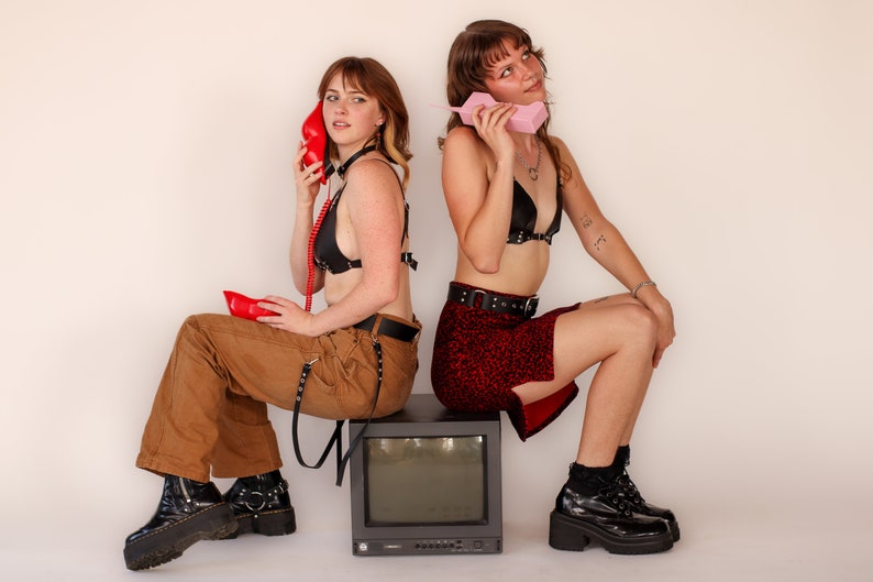 May include: Two people are seated, each holding a vintage telephone. One holds a red phone, the other a pink one. They are wearing black tops and boots. One wears brown pants, the other a red skirt. A vintage TV sits between them.
