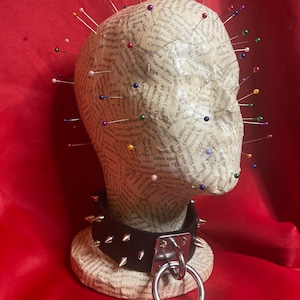 Può includere: Una testa di cartapesta ricoperta di spilli con un collare di pelle nera con punte argentate e un grande anello d'argento. La testa è su una base ricoperta di testo.