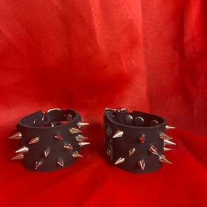 Puede incluir: Dos pulseras de cuero negro con pinchos plateados. Las pulseras están sobre un fondo rojo.