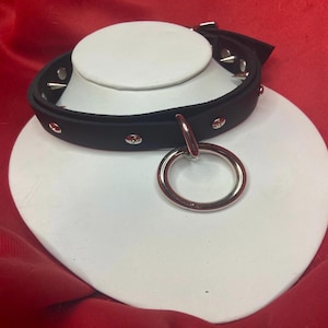 Può includere: Collana girocollo in pelle nera con borchie argentate e un grande ciondolo ad anello in metallo. La collana è esposta su una testa di manichino bianca su sfondo rosso. Chiusura con fibbia.