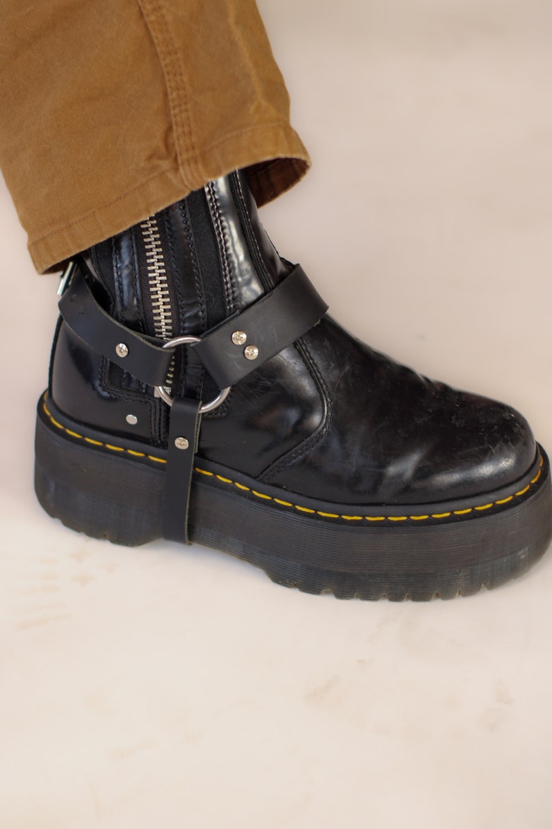 May include: Black platform boot with a silver zipper and a black leather harness featuring a silver ring and rivets. The boot has yellow stitching around the sole. A portion of brown pants is visible.