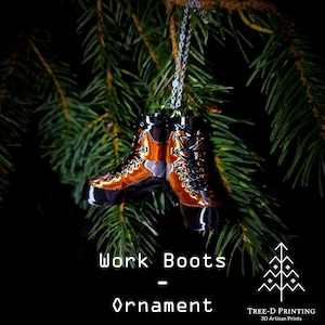 May include: A decorative ornament shaped like a pair of work boots, hanging from a silver chain against a backdrop of green pine needles. The boots are black, orange, and silver, with intricate detailing. The text "Work Boots - Ornament" is below the boots.