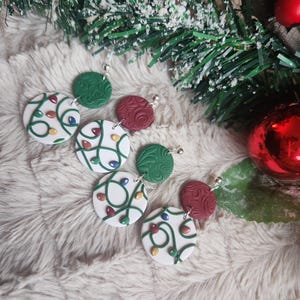 May include: A pair of Christmas-themed earrings with white, green, and red clay ornaments. Each ornament features a white background with green string lights and colorful bulbs.