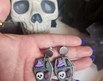 May include: A pair of Halloween-themed earrings. Each earring is shaped like a coffin and features a purple witch hat, books, a skull, and a pumpkin. The earrings are silver with a round silver stud.