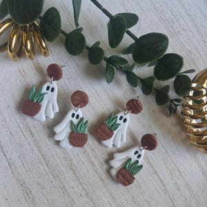 May include: Two pairs of dangle earrings featuring white ghost shapes holding small brown terracotta-colored pots with green plants. Each earring has a round brown stud top with a textured chevron pattern and a silver-toned metal post backing.